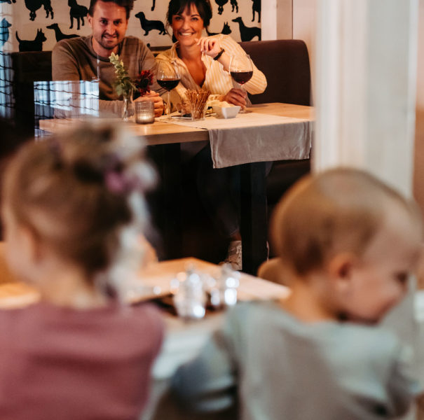 Ein Pärchen sitzt mit Rotwein im Restaurant und schaut lachend ihren zwei Kindern beim Spielen zu.