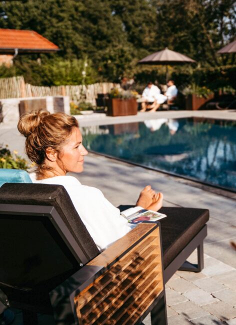 Blonde Frau sitzt in weißem Bademantel auf der Liege am Pool im Familienhotel Niedersachsen