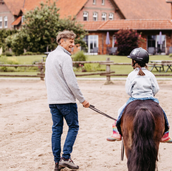 Opa führt seine Enkeltochter auf einem Reitplatz auf einem Pony.