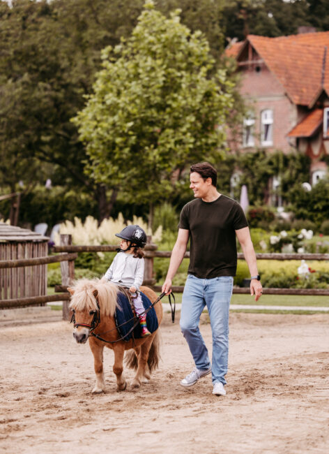 Ein Vater führt sein Kind auf dem Pony über den Reitplatz.